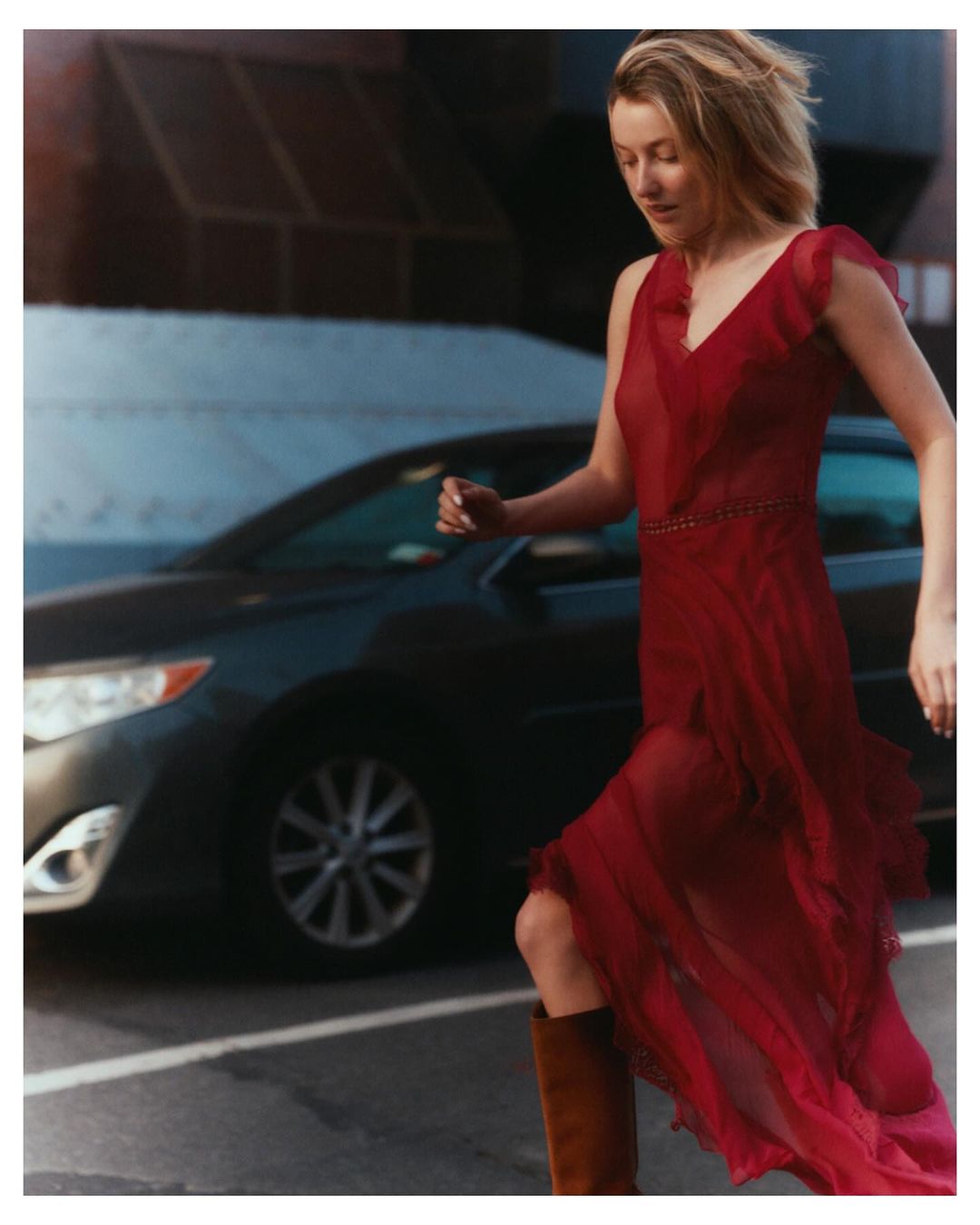Woman in a red dress strides across an urban street with a car in the background. She's wearing boots and has blonde hair.