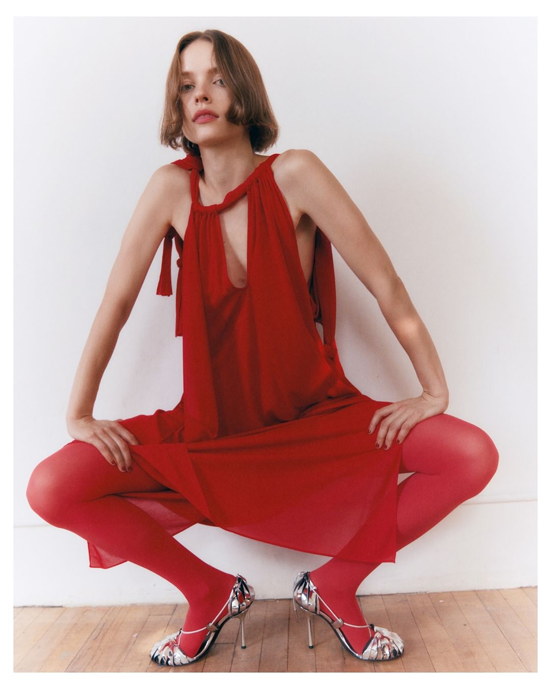 A woman in a red dress and tights poses indoors, showcasing a fashion-forward look with silver heels against a neutral background.