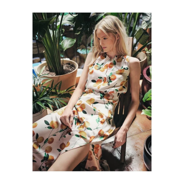 A blonde woman sits in a chair wearing an peach-printed sleeveless dress surrounded by potted plants in a well-lit indoor setting.