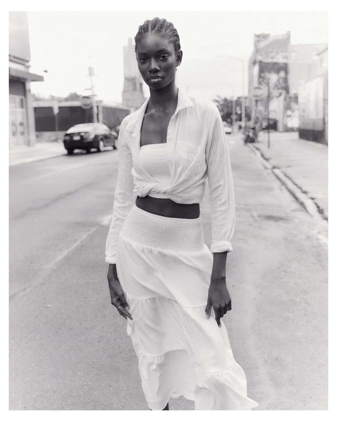 Monochrome portrait of an African-American woman in a white two-piece outfit on an urban street with a car in the background.
