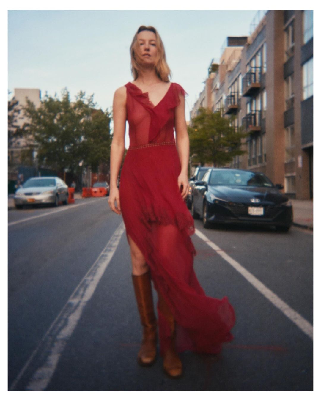 Woman in a red dress and brown boots walks down an empty city street with cars and buildings in the background on a sunny day.