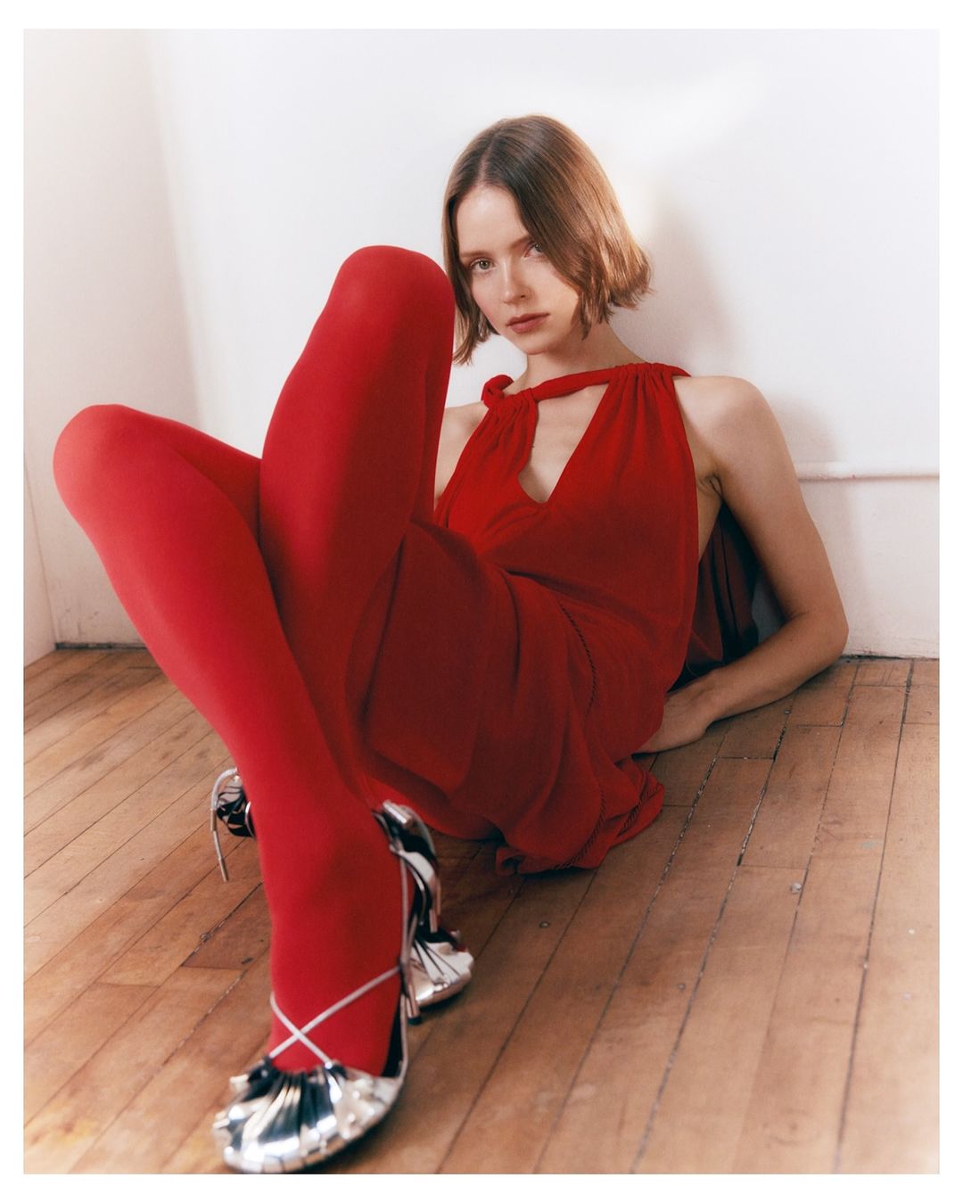 Young woman in a red dress and stockings sits on the wooden floor, posing with silver heels against a white wall backdrop indoors.