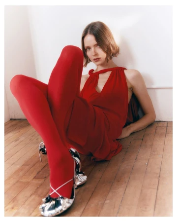 Young woman in a red dress and stockings sits on the wooden floor, posing with silver heels against a white wall backdrop indoors.