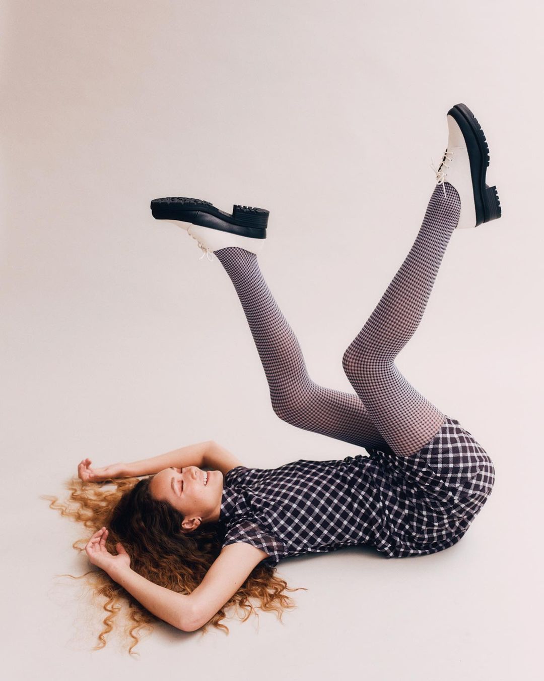 A smiling young woman with curly hair in a black and white checked dress and patterned tights is lying on her back with legs in the air.