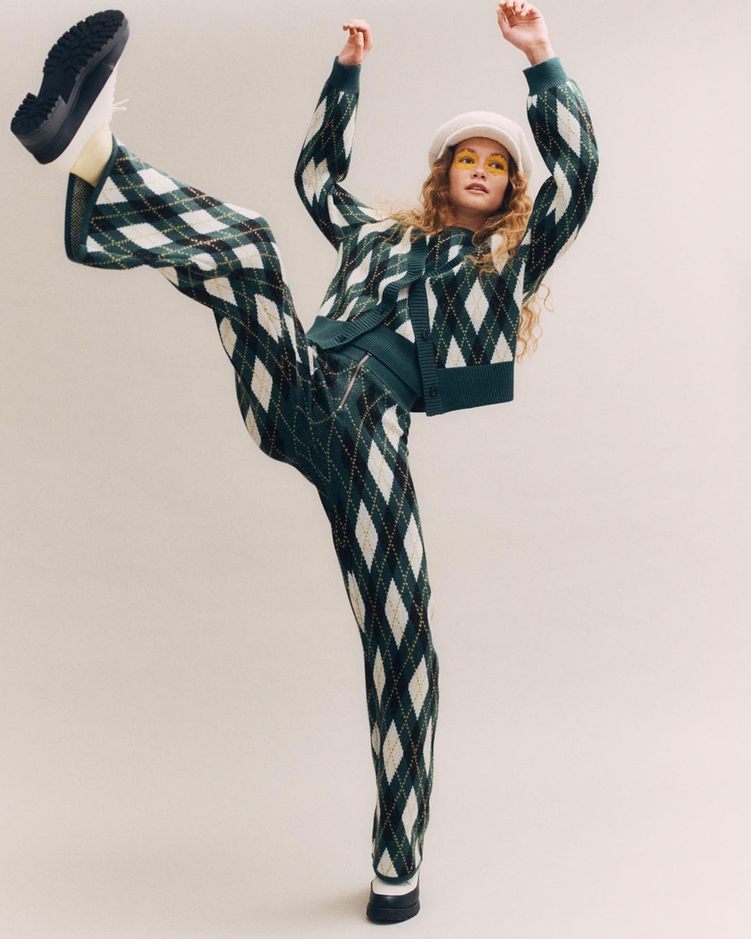 A slender, light-skinned woman in a green argyle knit cardigan and matching pants strikes a dynamic pose against a pale backdrop.