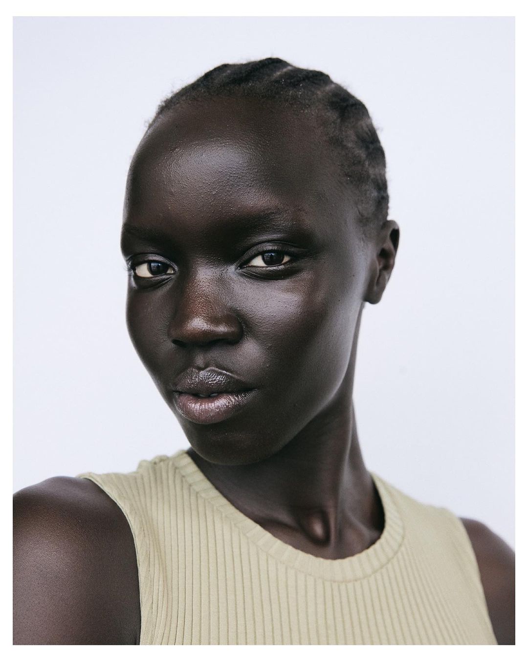 Close-up portrait of a woman with dark skin and braided hair, wearing a light-green ribbed tank top against a white background.