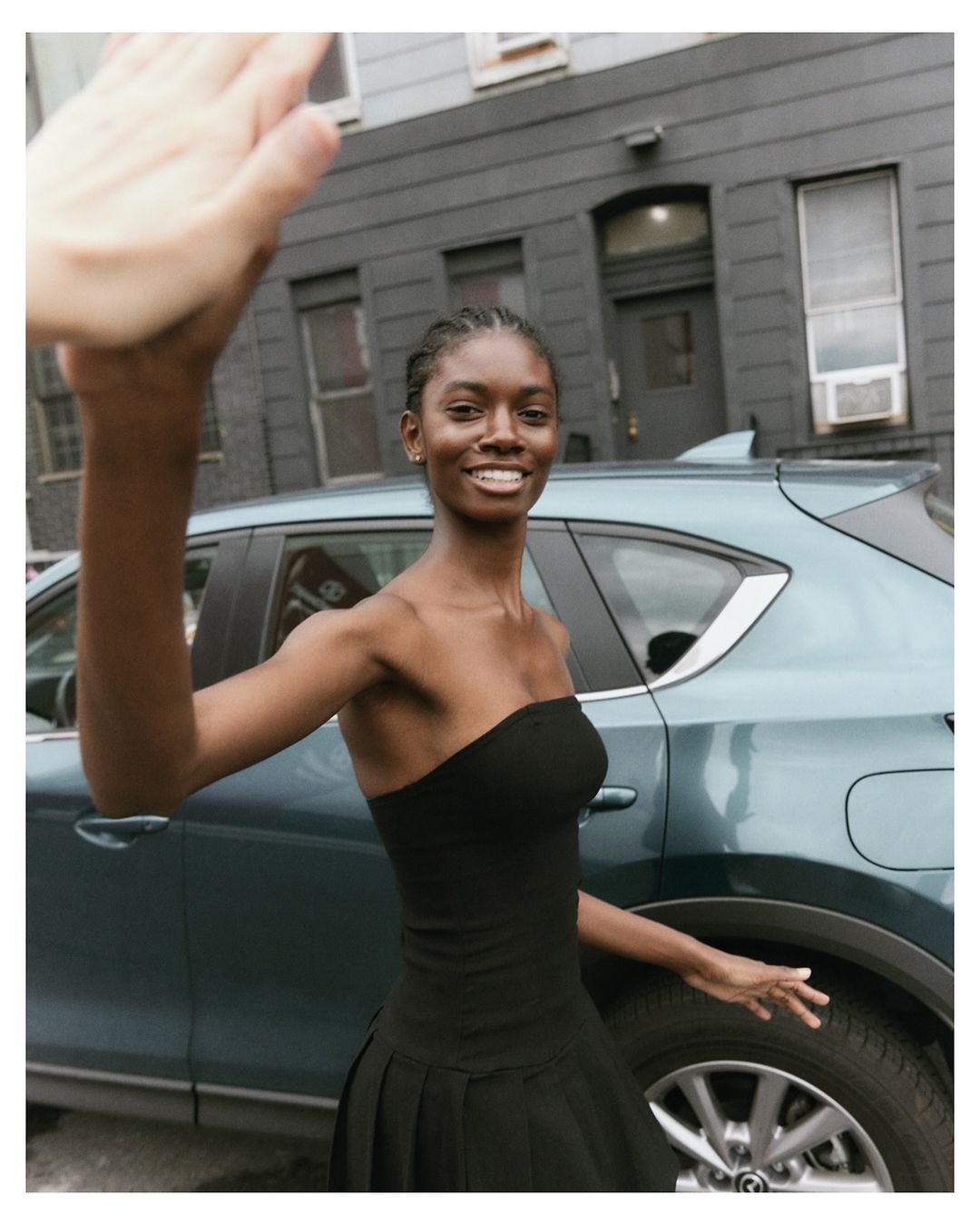 A smiling black woman in a black dress waves in front of a teal car in an urban setting, creating a stylish and candid moment.