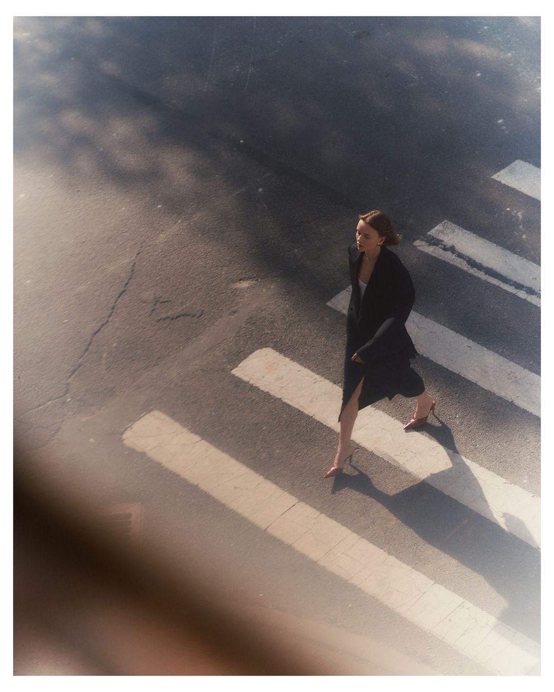 A woman walks across a crosswalk wearing a dark coat and light-colored heels in full sunlight, shot from a high angle in an urban environment.