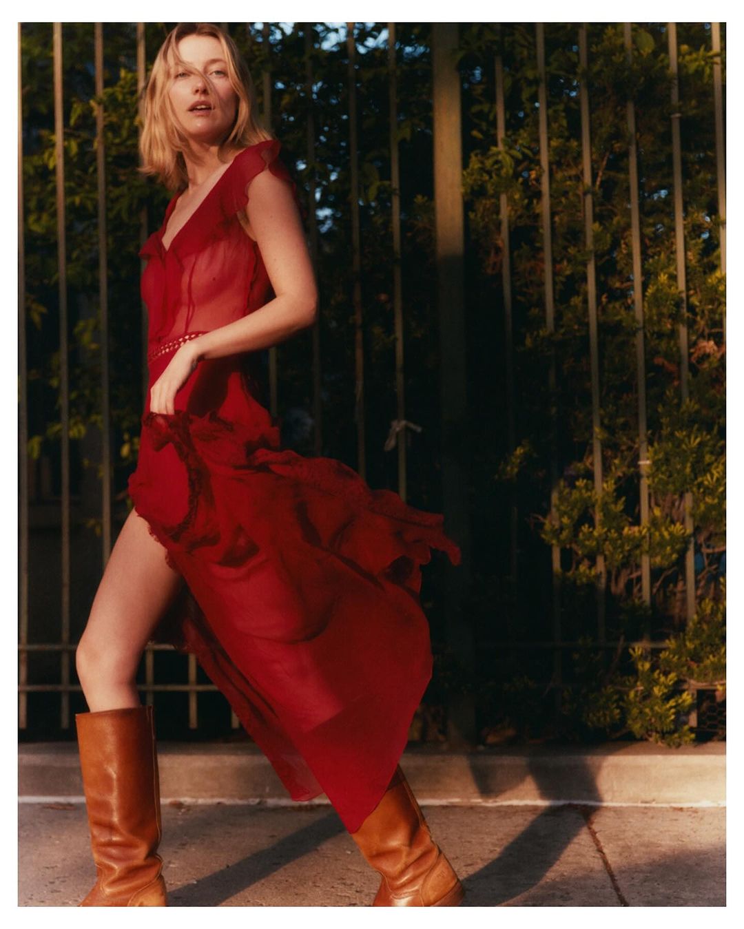 A woman in a red dress and brown boots poses outdoors against a dark leafy fence, bathed in warm sunlight, creating a fashion editorial image.