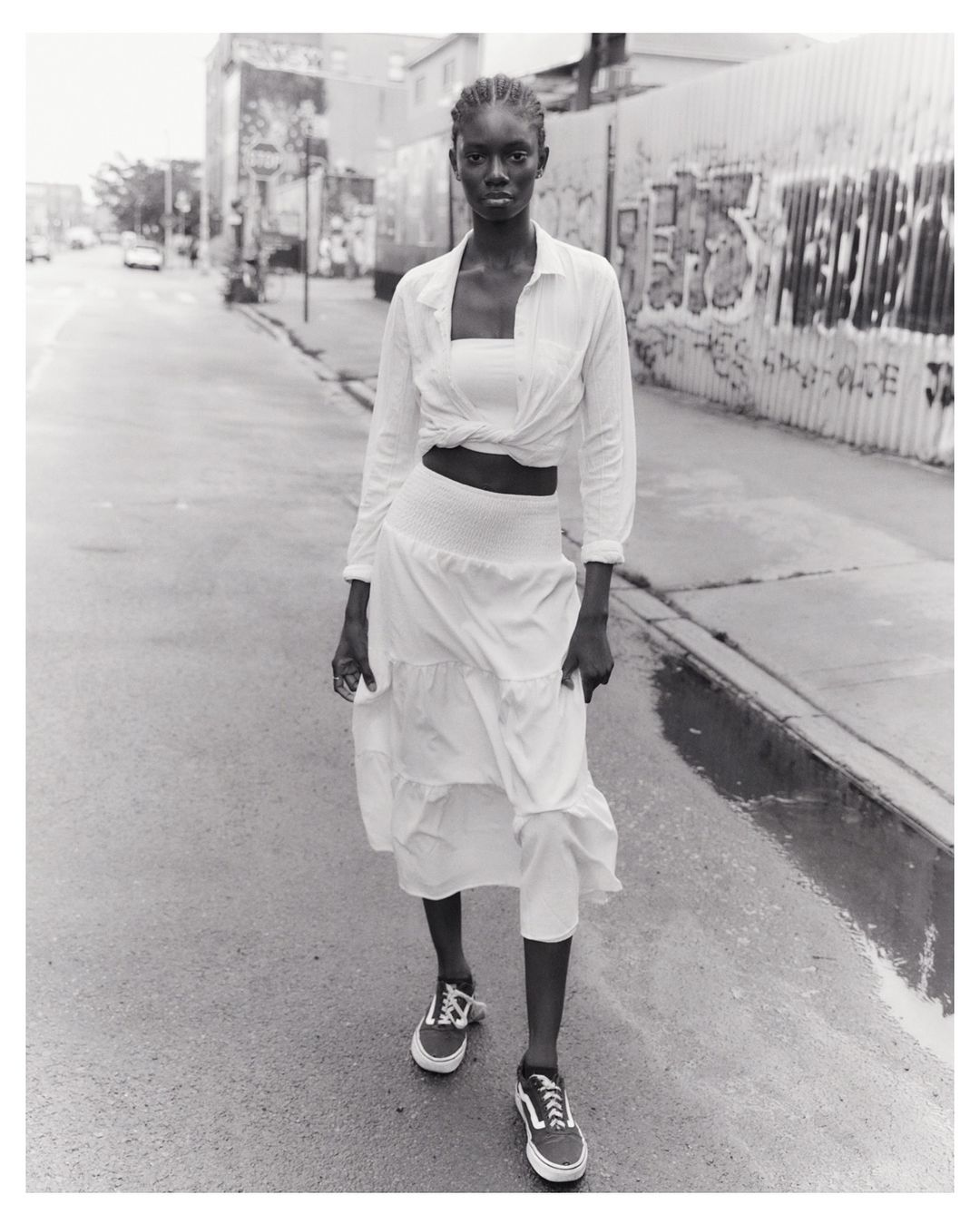 Monochrome portrait of a Black woman walking on a city street, wearing a white two-piece outfit and sneakers, with graffiti in background.