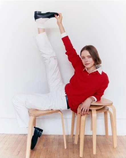 Fashion shot of woman in red top and white pants seated on wooden stools holding black shoe overhead in a white studio.