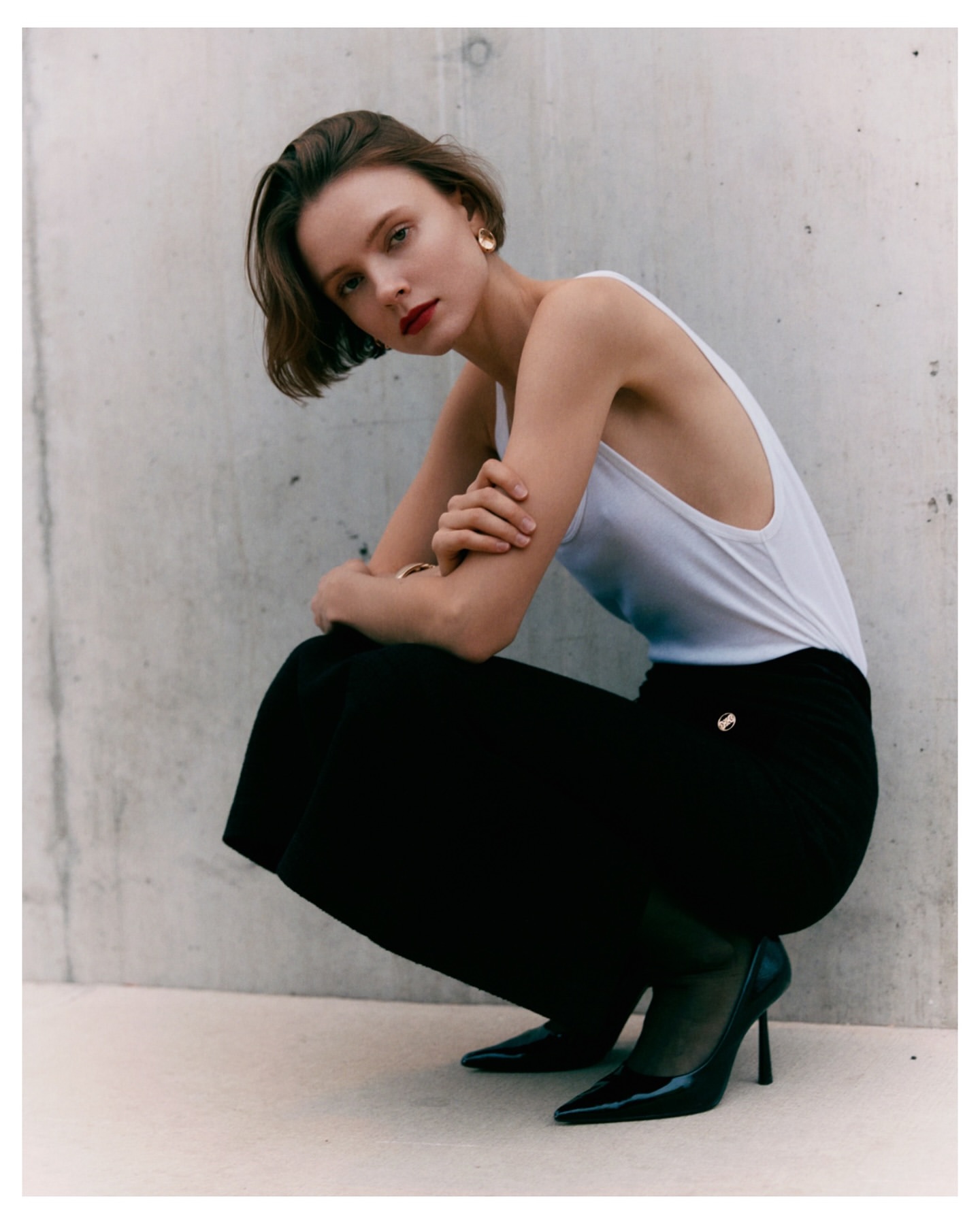 Young woman in white top and black pants poses in an urban setting with high heels. The background is a concrete wall.