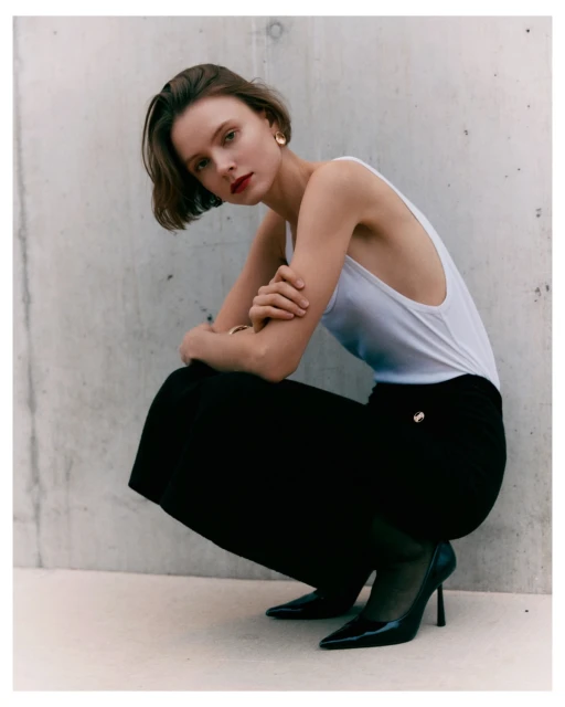 Young woman in white top and black pants poses in an urban setting with high heels. The background is a concrete wall.