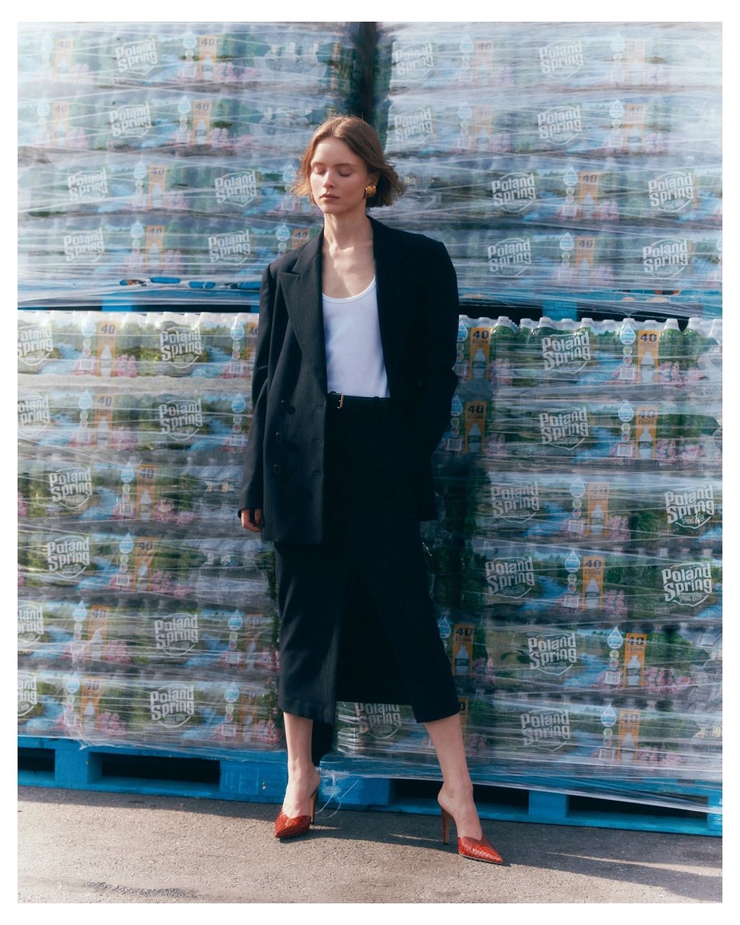 Fashion shot of a female model in an oversized blazer, skirt, and heels, posed in front of a stack of Poland Spring water bottles.