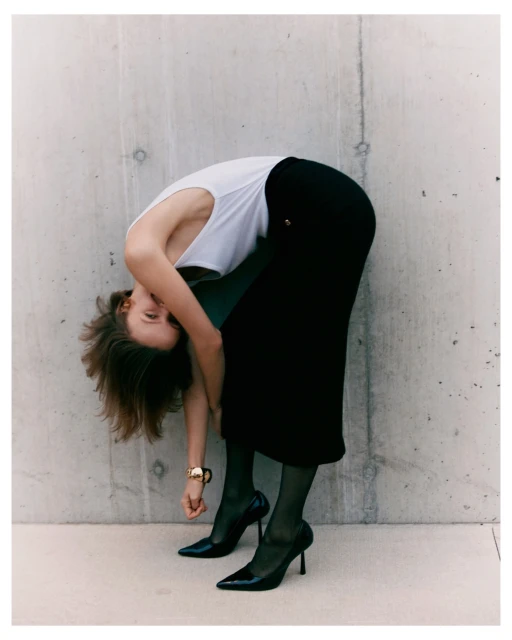Adult woman in a white top and black skirt bending over with shiny black heels in front of a grey concrete wall, studio lighting.