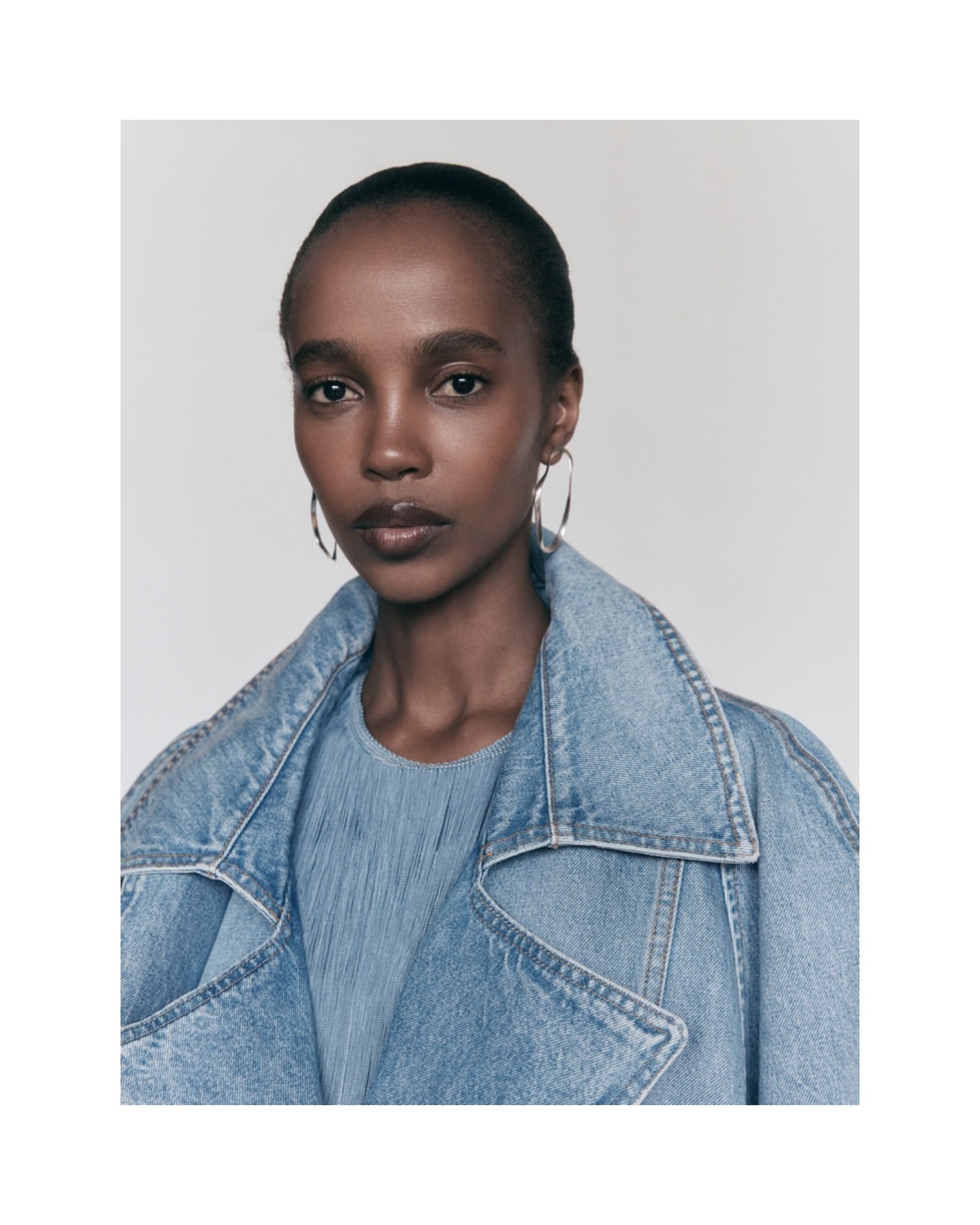 Close-up portrait of a young, Black woman in a denim coat, wearing silver hoop earrings, against a neutral grey background in soft lighting.
