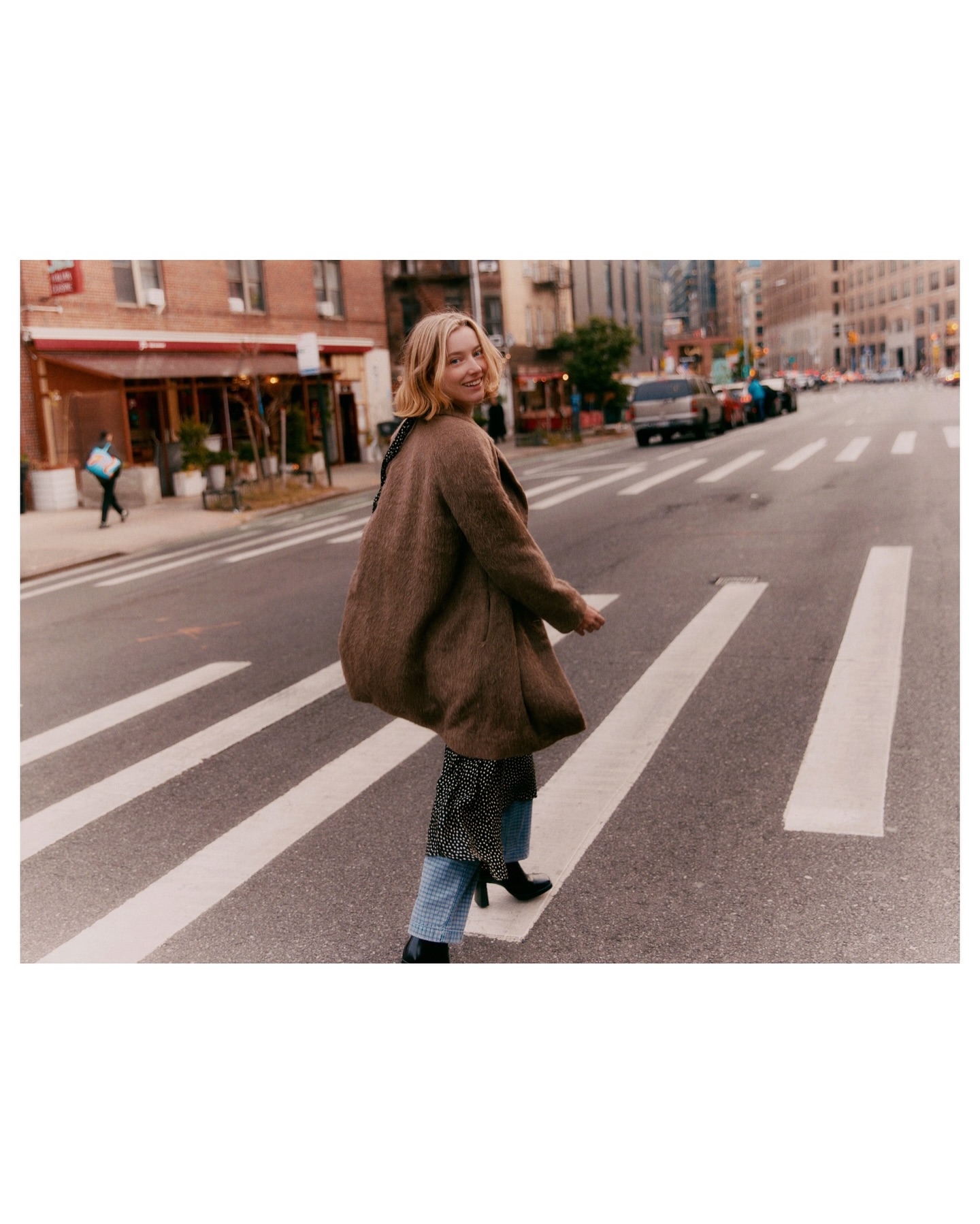 Woman in a brown coat walks across a zebra crossing in a city street. Buildings and parked cars are in the background.