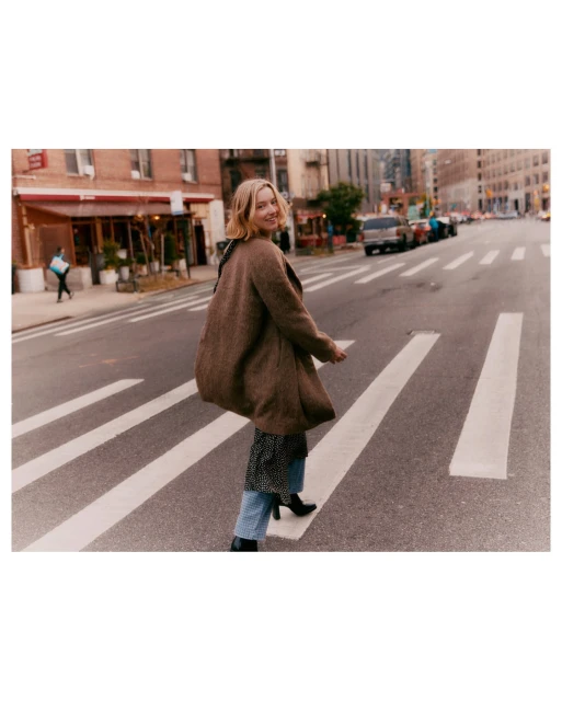 Woman in a brown coat walks across a zebra crossing in a city street. Buildings and parked cars are in the background.