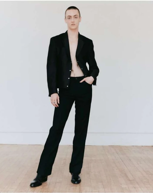 Adult fashion model poses indoors wearing an open black jacket, black pants, and black shoes against a white wall and light wood floor.