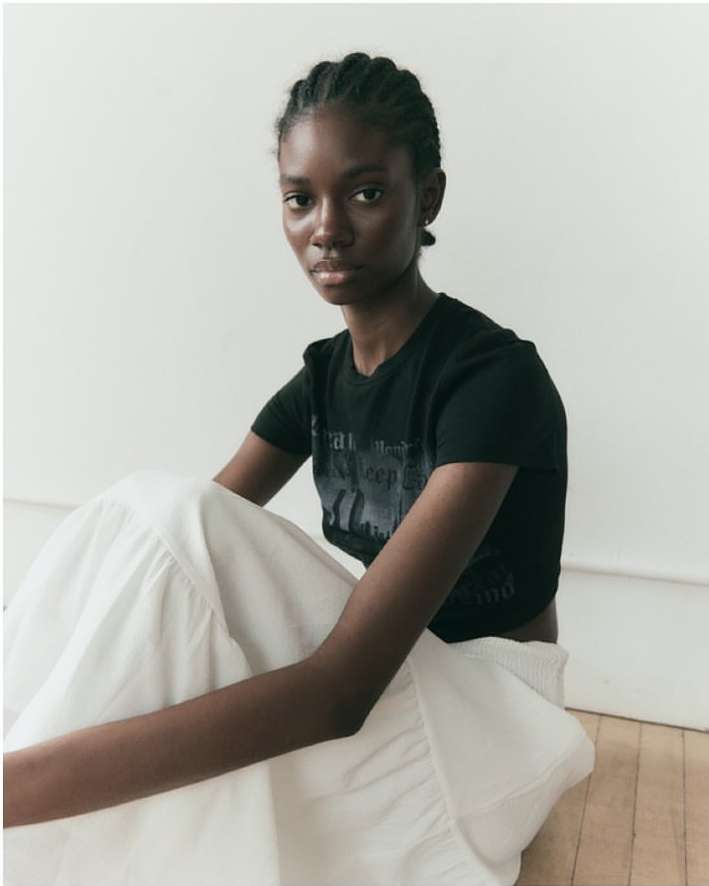 Portrait of an African American woman in a black tee shirt and large white skirt sitting against a white wall in a photo studio.