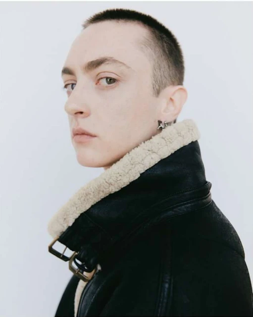 Portrait of a person with a shaved head, gazing over their shoulder, wearing a black leather aviator jacket with cream-colored fur trim.