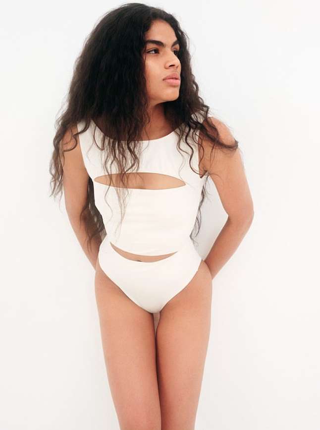 Young woman with long dark hair poses in a white one-piece outfit against a white background. The outfit features cut-out details.