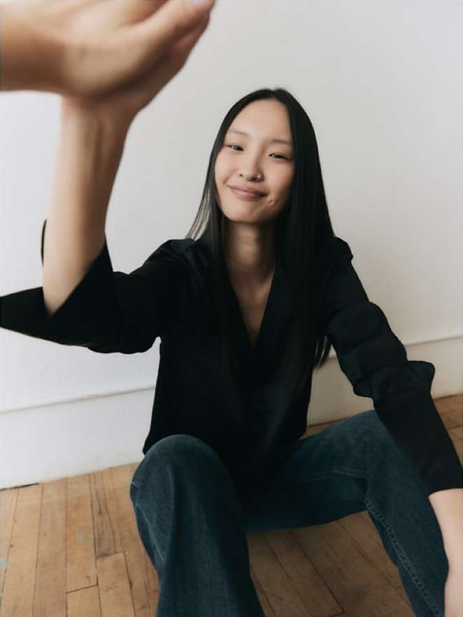 Smiling Asian woman in black shirt and blue jeans sitting on wooden floor with arm raised indoors against white wall.