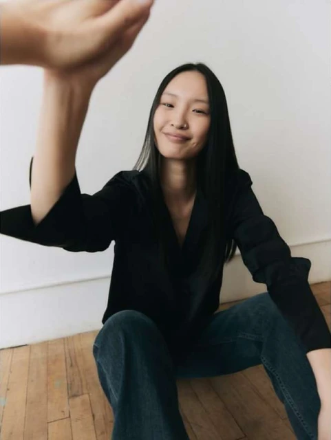 Smiling Asian woman in black shirt and blue jeans sitting on wooden floor with arm raised indoors against white wall.