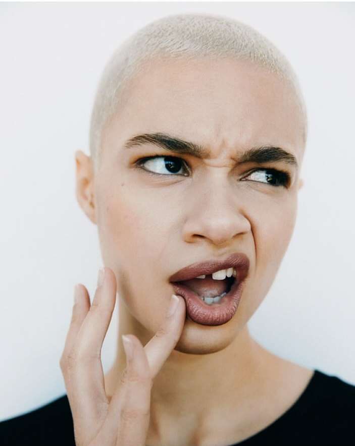 Close-up portrait of a woman with short platinum blonde hair, making a face and touching her lips against a plain white background.