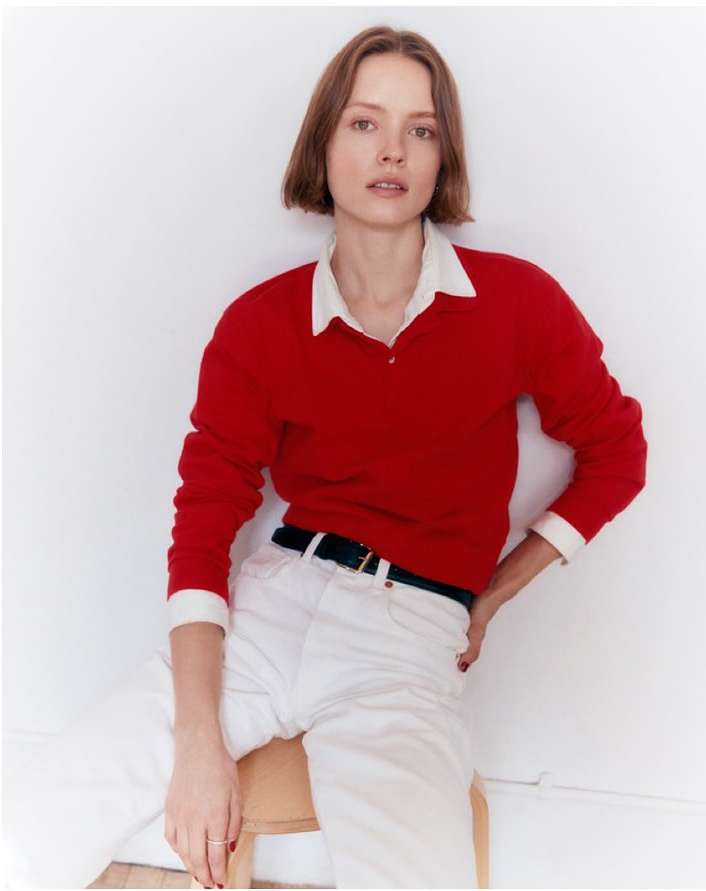 Portrait of a fair-skinned adult female in a red sweater, white collared shirt, and white jeans, sitting on a wooden stool against a white wall.