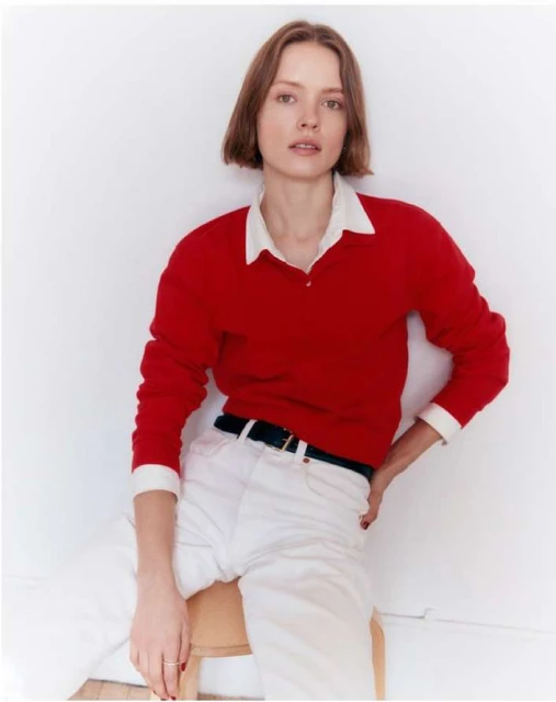 Portrait of a fair-skinned adult female in a red sweater, white collared shirt, and white jeans, sitting on a wooden stool against a white wall.