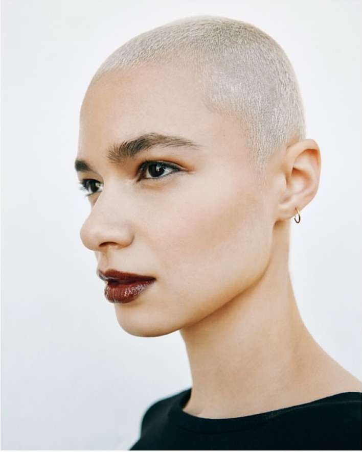 Close-up studio portrait of a fair-skinned adult woman with a buzzcut, wearing dark lipstick, jewelry and a black top against a white background.