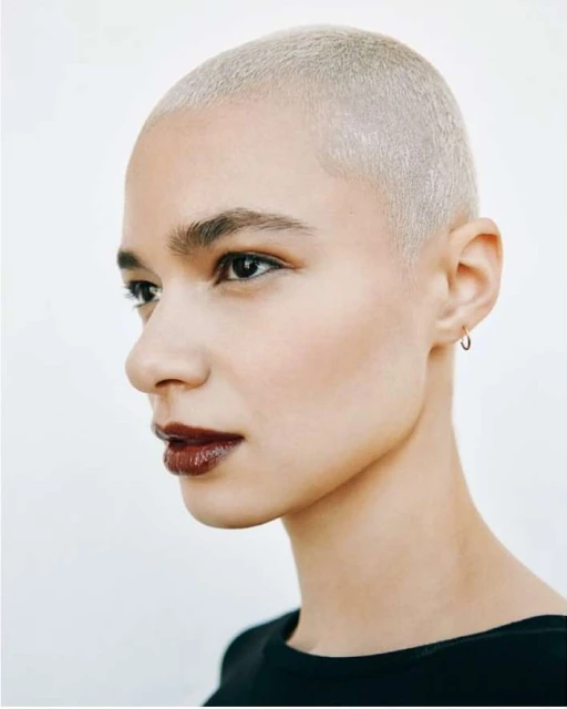 Close-up studio portrait of a fair-skinned adult woman with a buzzcut, wearing dark lipstick, jewelry and a black top against a white background.