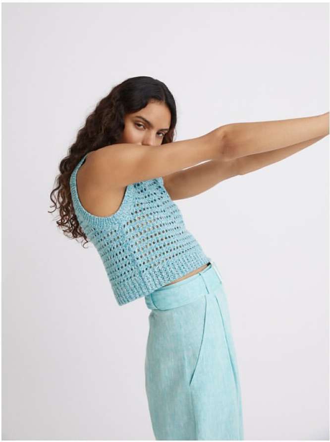 A young woman poses wearing a blue knit top and light blue slacks against a light background in a fashion shot with her arms raised.