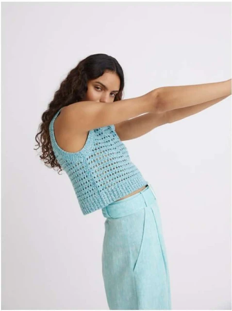 A young woman poses wearing a blue knit top and light blue slacks against a light background in a fashion shot with her arms raised.