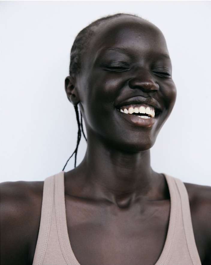 Portrait of a Black woman smiling with a tan tank top against a white background in a studio with eye-level camera angle.