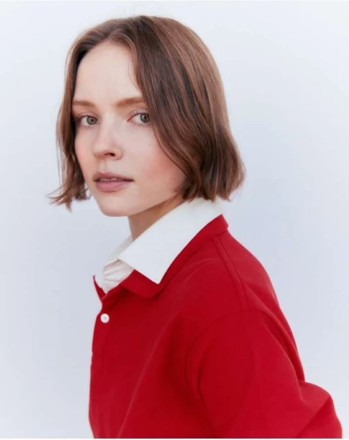 Close-up of a young fair-skinned woman in a red collared shirt, set against a neutral background in a studio lighting setup.