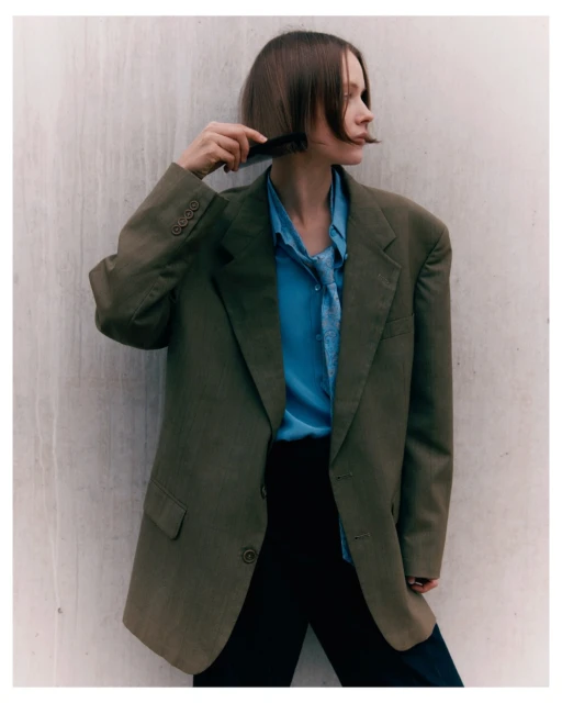A young adult woman with brown hair poses in a bright, neutral setting, wearing an oversized blazer with a blue top and holding a comb.