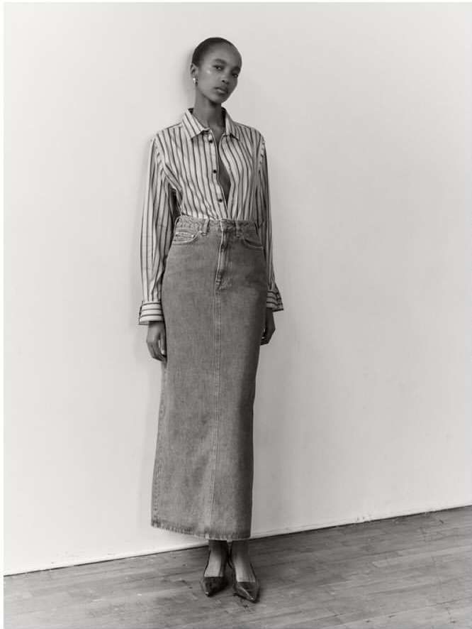 Black and white editorial portrait of a woman wearing a striped button-down shirt and long denim skirt against a plain wall.