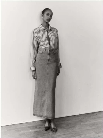 Black and white editorial portrait of a woman wearing a striped button-down shirt and long denim skirt against a plain wall.