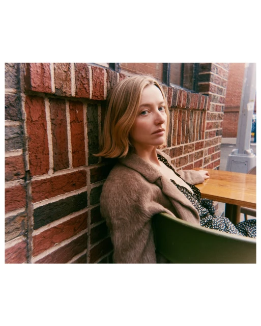Adult woman poses outdoors in a speckled dress and tan coat against a brick wall with a wooden table and green chair.