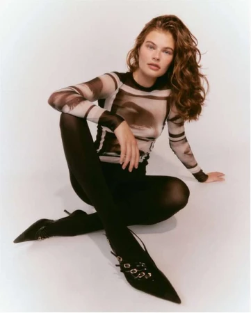 Model with long brown hair wearing a printed long-sleeve top and black tights seated against a white background