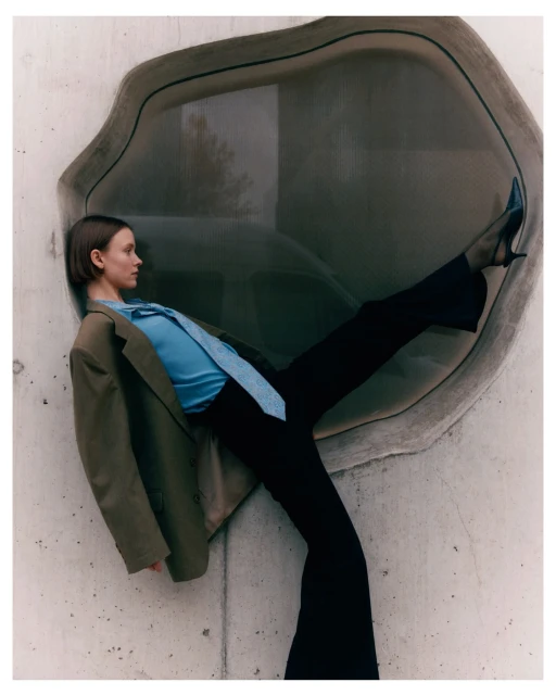 Woman in 70s-inspired fashion, olive blazer, blue top, black pants, blue heels, posing against concrete wall with round window.