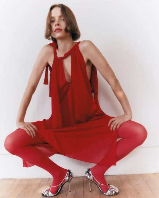 A slender woman in a red dress and tights strikes a squatting pose on a wood floor against a white wall, accentuated by high-heeled sandals.