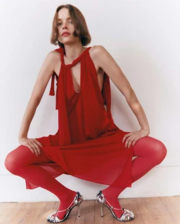 A slender woman in a red dress and tights strikes a squatting pose on a wood floor against a white wall, accentuated by high-heeled sandals.
