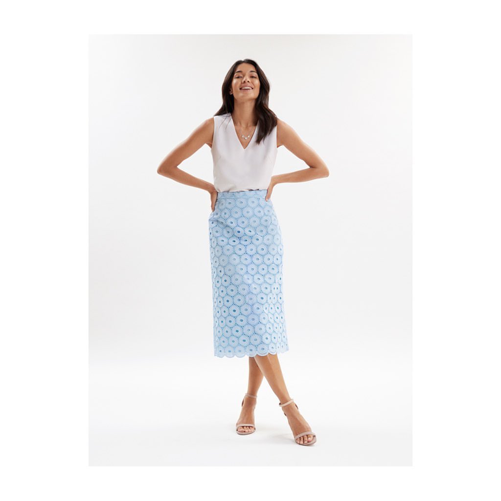 Smiling woman posing in a white V-neck top and light blue eyelet pencil skirt with nude ankle-strap heels in a bright studio.