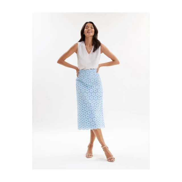 Smiling woman posing in a white V-neck top and light blue eyelet pencil skirt with nude ankle-strap heels in a bright studio.