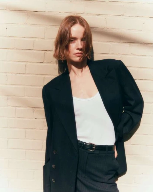 An adult woman with auburn hair poses in front of a brick wall wearing a blazer, white top, dark pants and a belt, captured in medium shot.