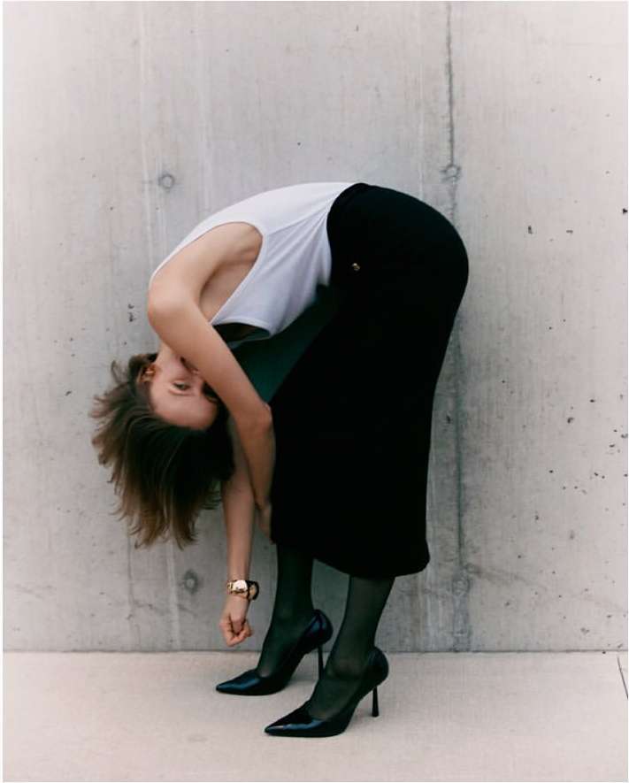 Adult female bending over, wearing a white tank top, black skirt, black sheer tights, and black high heels against a concrete wall.