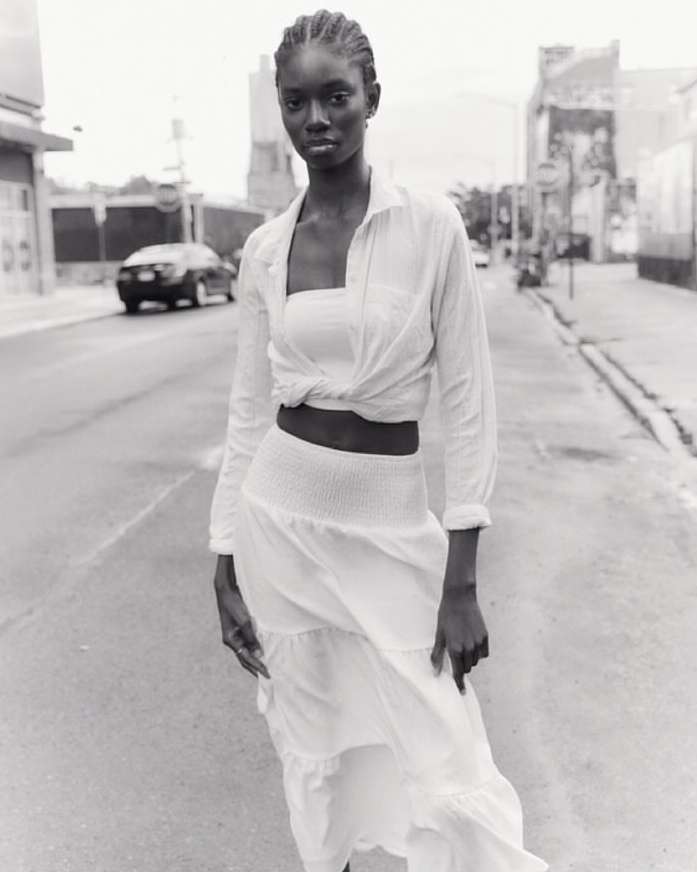 Black and white editorial portrait of a woman standing on a city street wearing a light-coloured skirt and knotted shirt.